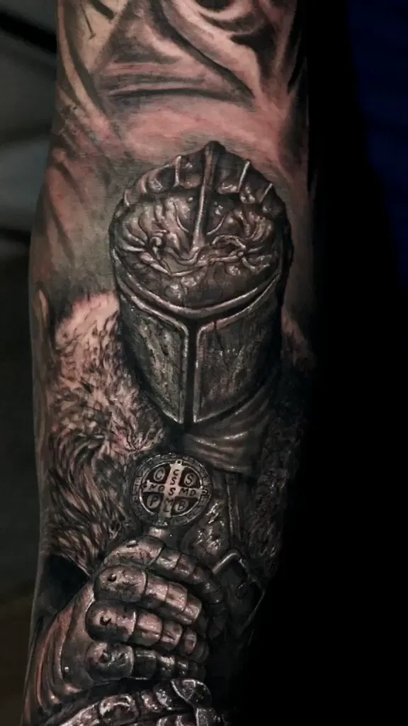 Tatuagem realista de cavaleiro de armadura segurando a medalha de São Bento, no antebraço