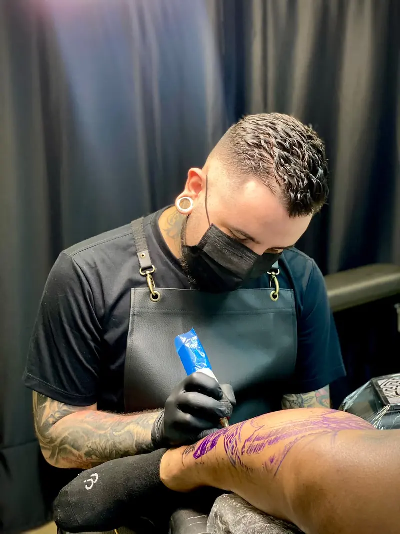 Mikayo Kelmer de máscara e luvas tatuando a perna de um cliente