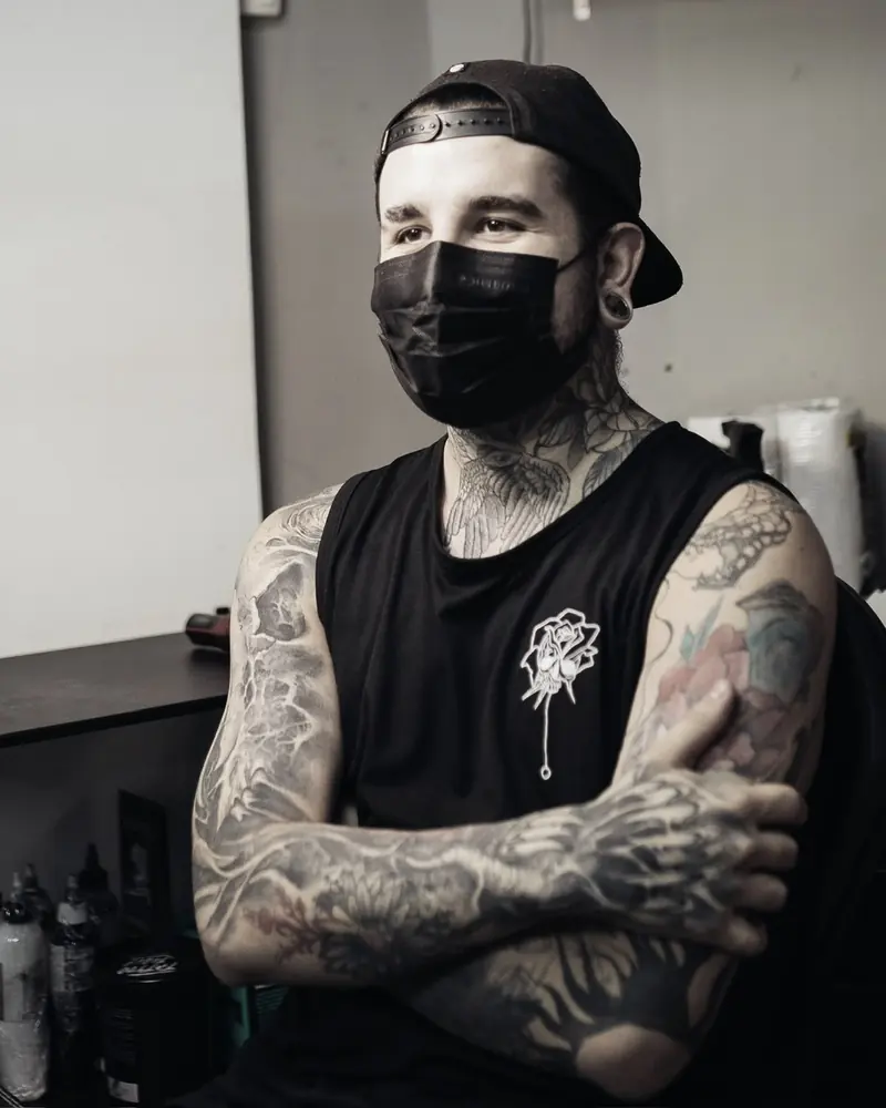 Retrato em preto e branco de Mikayo Kelmer, de boné e máscara, braços tatuados cruzados