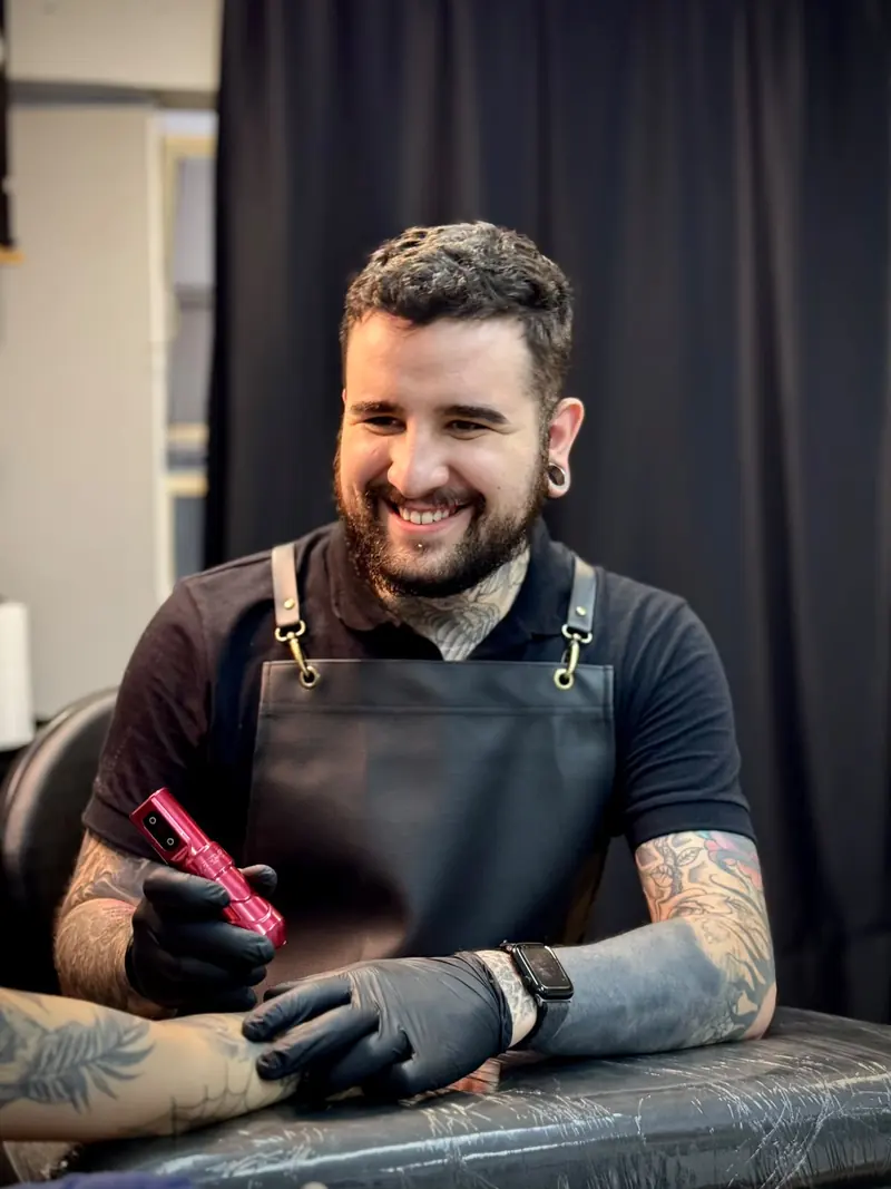 Mikayo Kelmer sorrindo com a máquina de tatuagem na mão, de avental preto